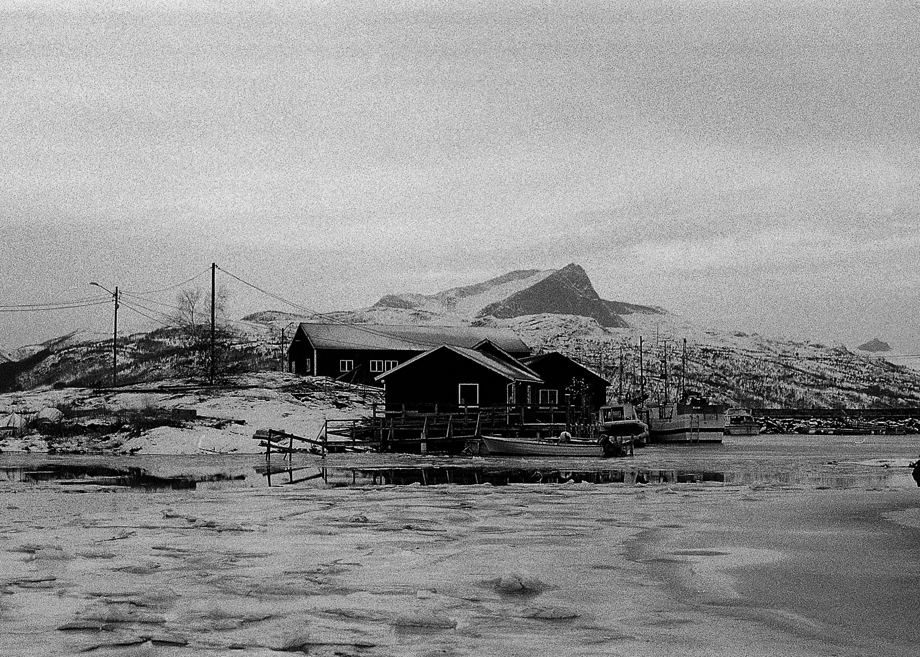 A black and white photograph of a boat house and some boats in a Strøksnes harbour in winter time. Ice is on the water and there is a snow covered mountain in the distance.